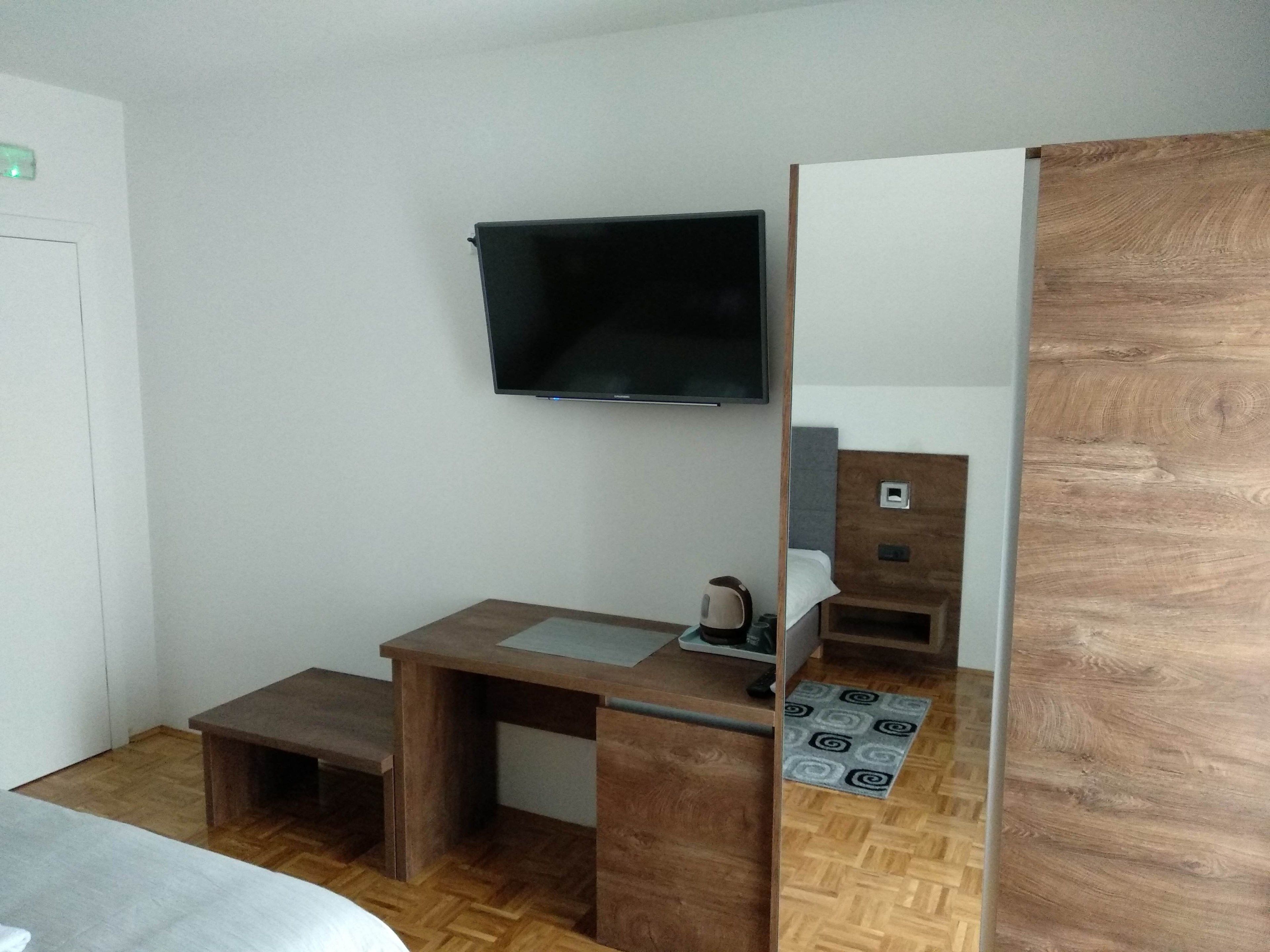 A compact hotel room with a wooden desk, a flat-screen TV mounted on the wall, a small bedside table, and a bed with a white mattress.