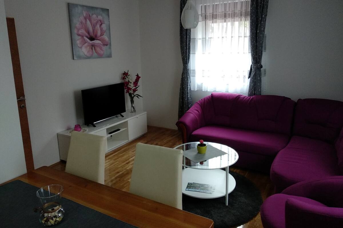 A living room with a TV on a white stand, a purple sofa, a glass coffee table, and a painting of a flower on the wall.