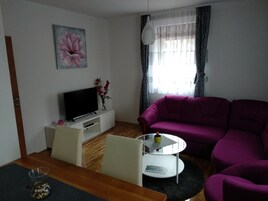 Una sala de estar con un televisor sobre un mueble blanco, un sofá morado, una mesa de centro de vidrio y un cuadro de una flor en la pared.