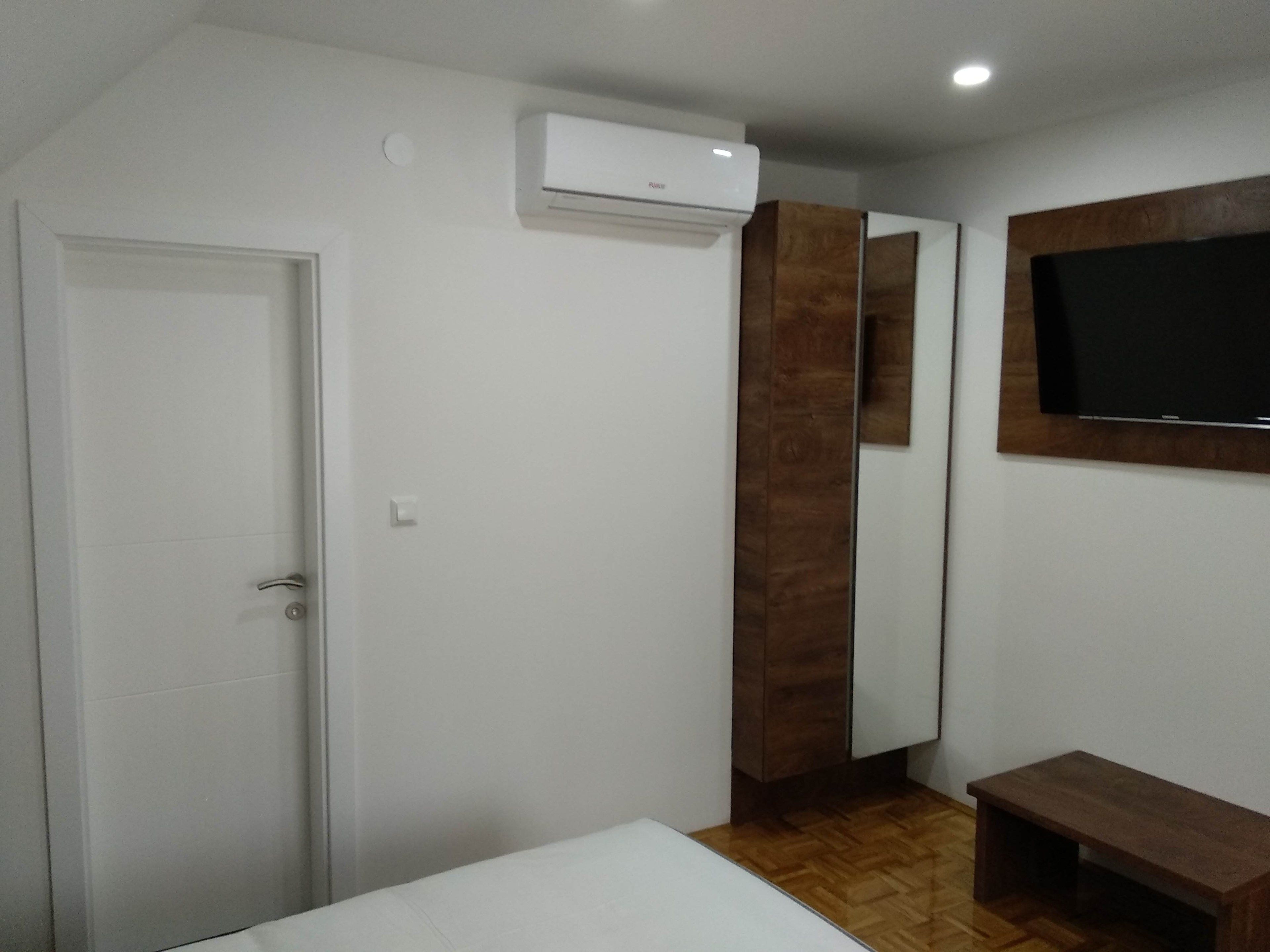 A bedroom with a bed, a TV mounted on the wall, a wardrobe, and an air conditioning unit.