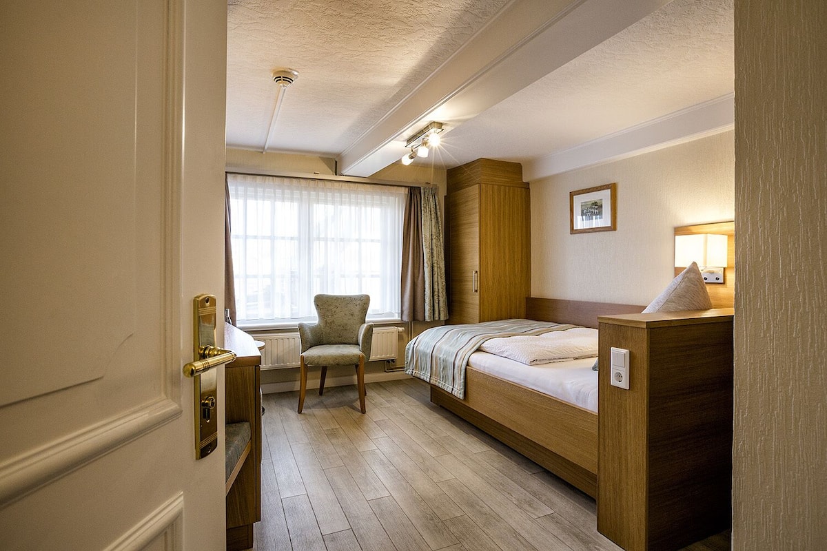 Una habitación de hotel con cama, una silla, escritorio y ventana con cortinas.