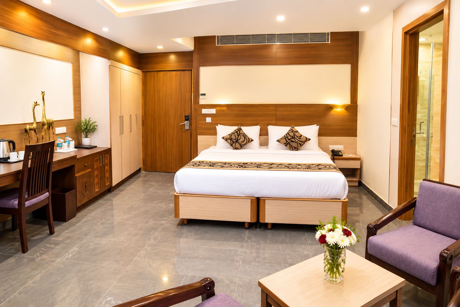 A hotel room with a bed, desk, chair, and a small table with a vase of flowers.