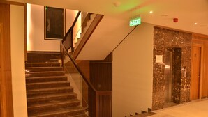 A staircase with a wooden handrail and a glass railing, leading to an elevator. - Hotel Placid (Prayagraj)