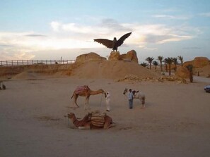 A desert landscape with a camel, a person on a camel, and a large bird statue. - Résidence El Arich (Tozeur)