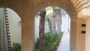 A corridor with brick arches, a tiled floor, and a pathway lined with plants and palm trees. - Résidence El Arich (Tozeur)