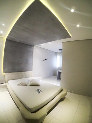 A modern bedroom with a curved ceiling, a bed with a logo, a small bedside table, and a nightstand. - Motel Monza (Santos)