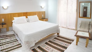 Un dormitorio ordenado con cabecera de madera, colcha blanca, alfombra estampada, mesita de noche pequeña y una silla.