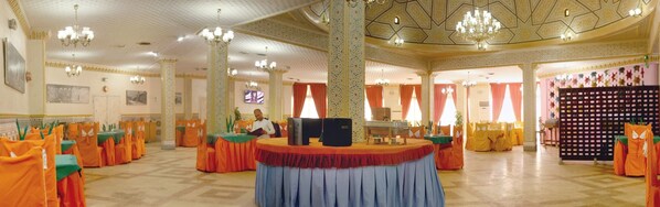 A spacious hall with ornate ceiling and chandeliers, tables with orange tablecloths, and a central stage with a sound system.
