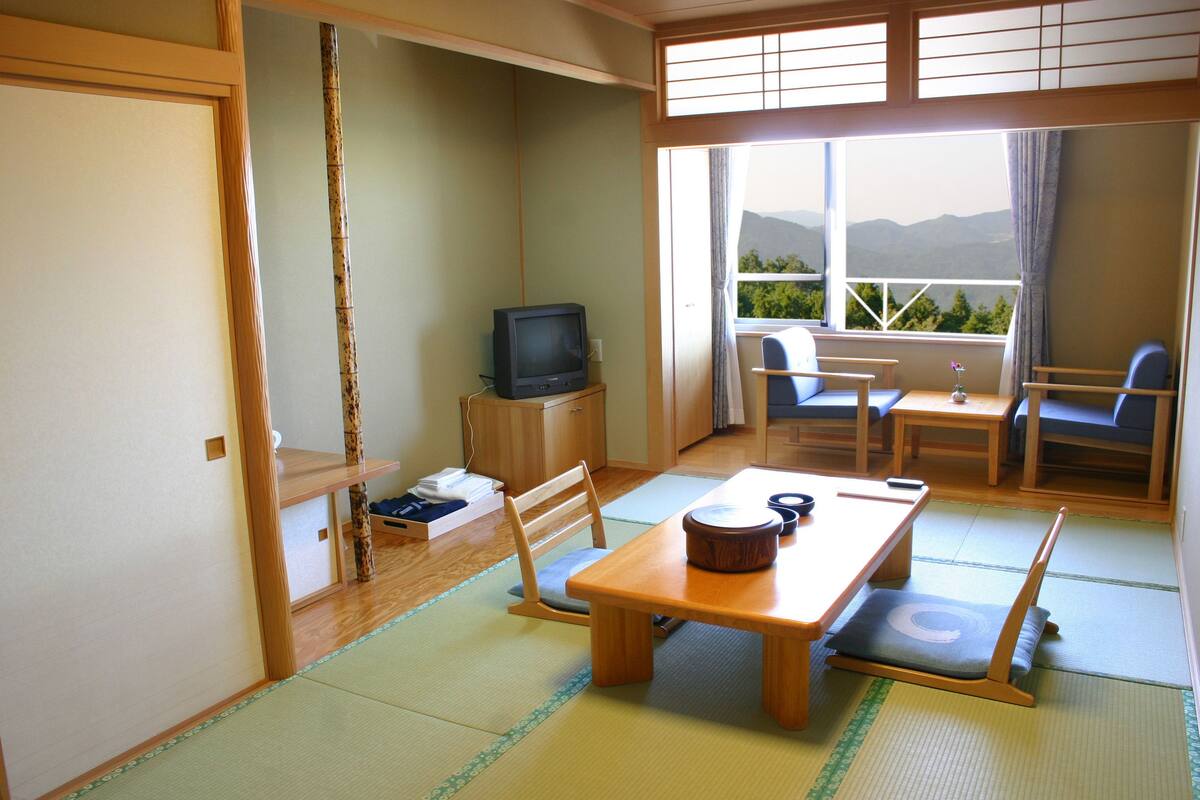 Una habitación japonesa tradicional con suelo de tatami, una mesa de madera baja, un televisor y vistas a montañas por la ventana.