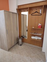 Un armario de madera con espejo, un pequeño cubo de basura y una estantería con una caja de pañuelos y revistas.
