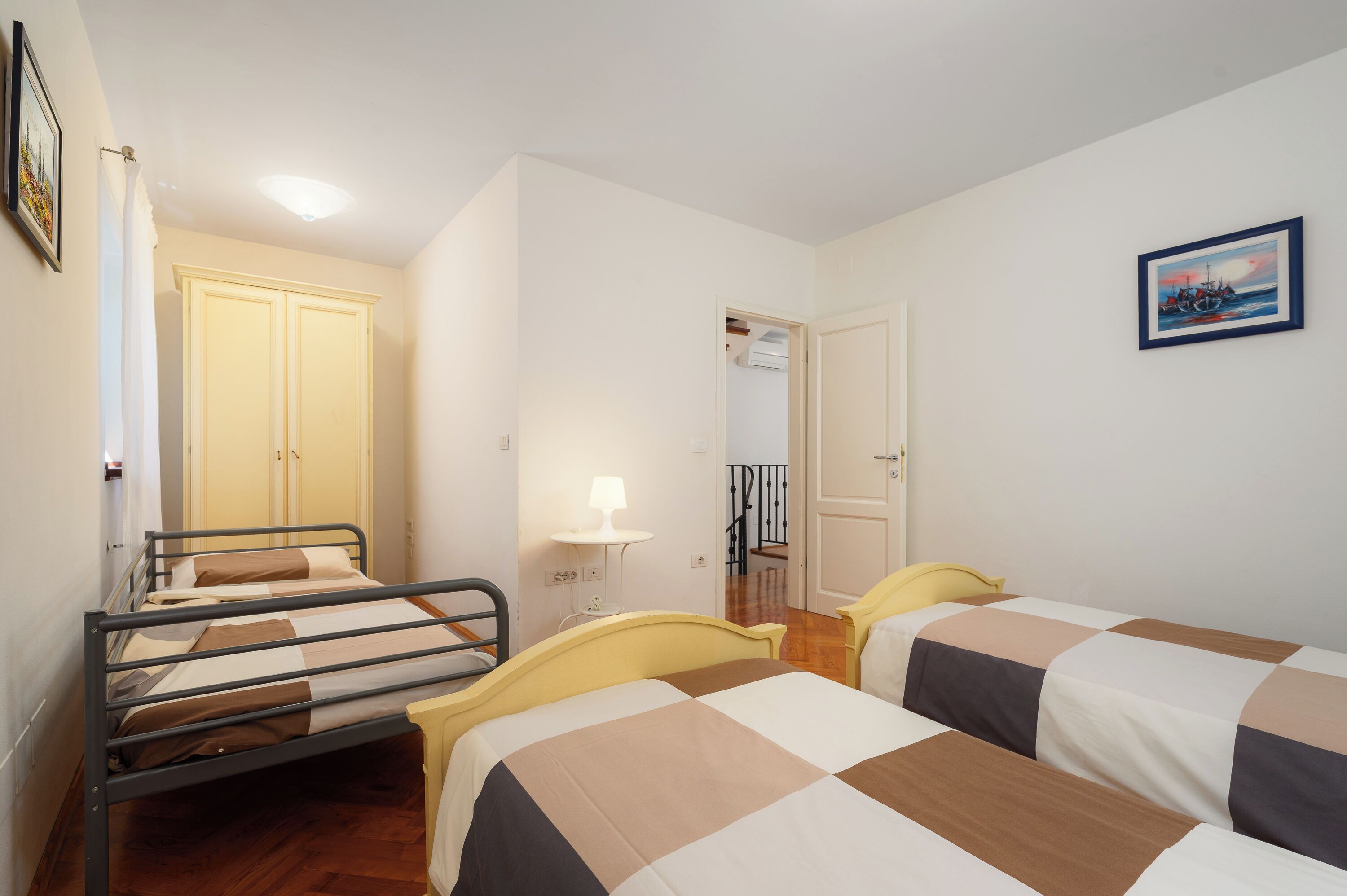 A room with two beds, a small table with a lamp, a wardrobe, and a painting on the wall.