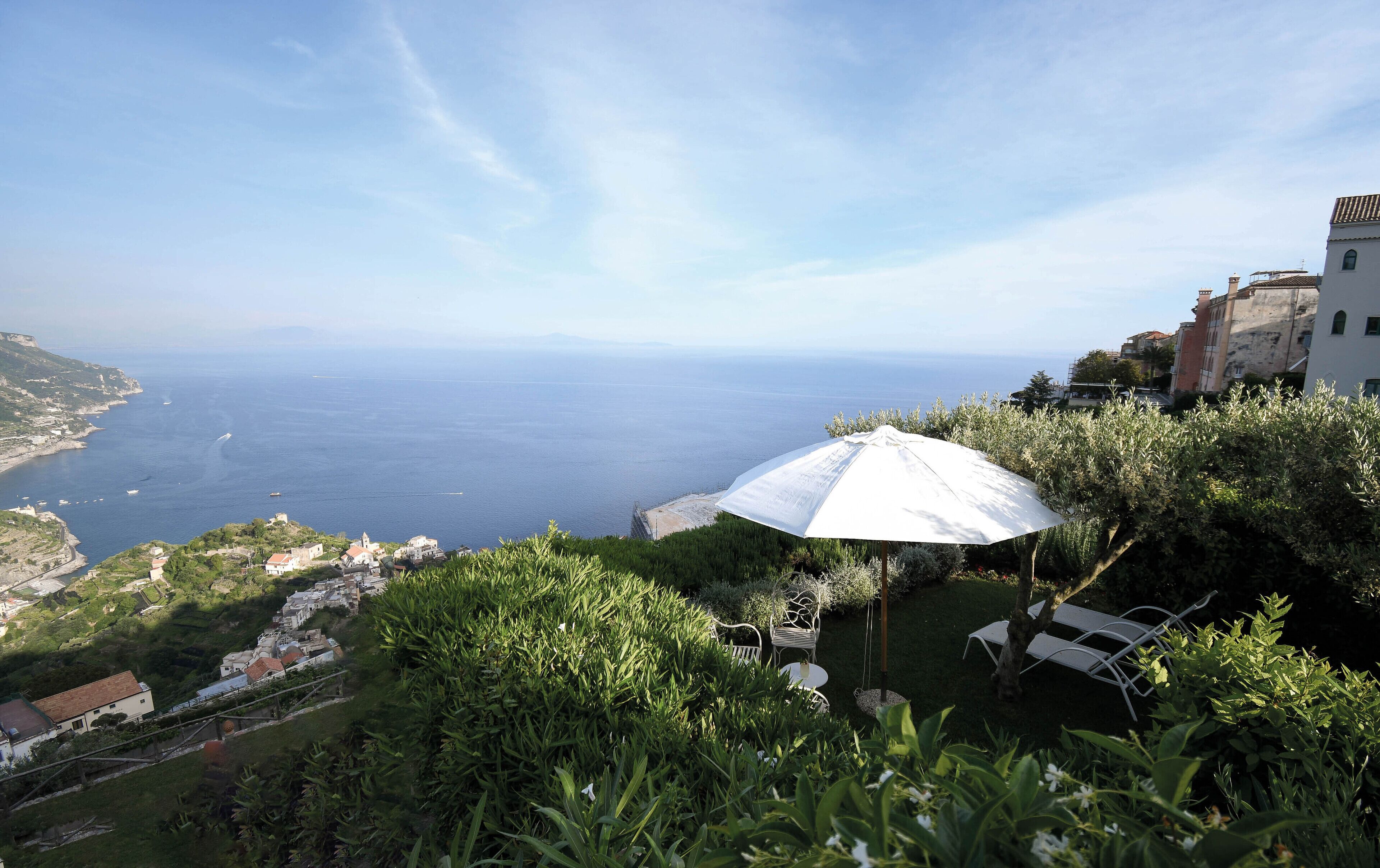 Vista sulla costa con un ombrellone bianco, vegetazione e edifici sullo sfondo.