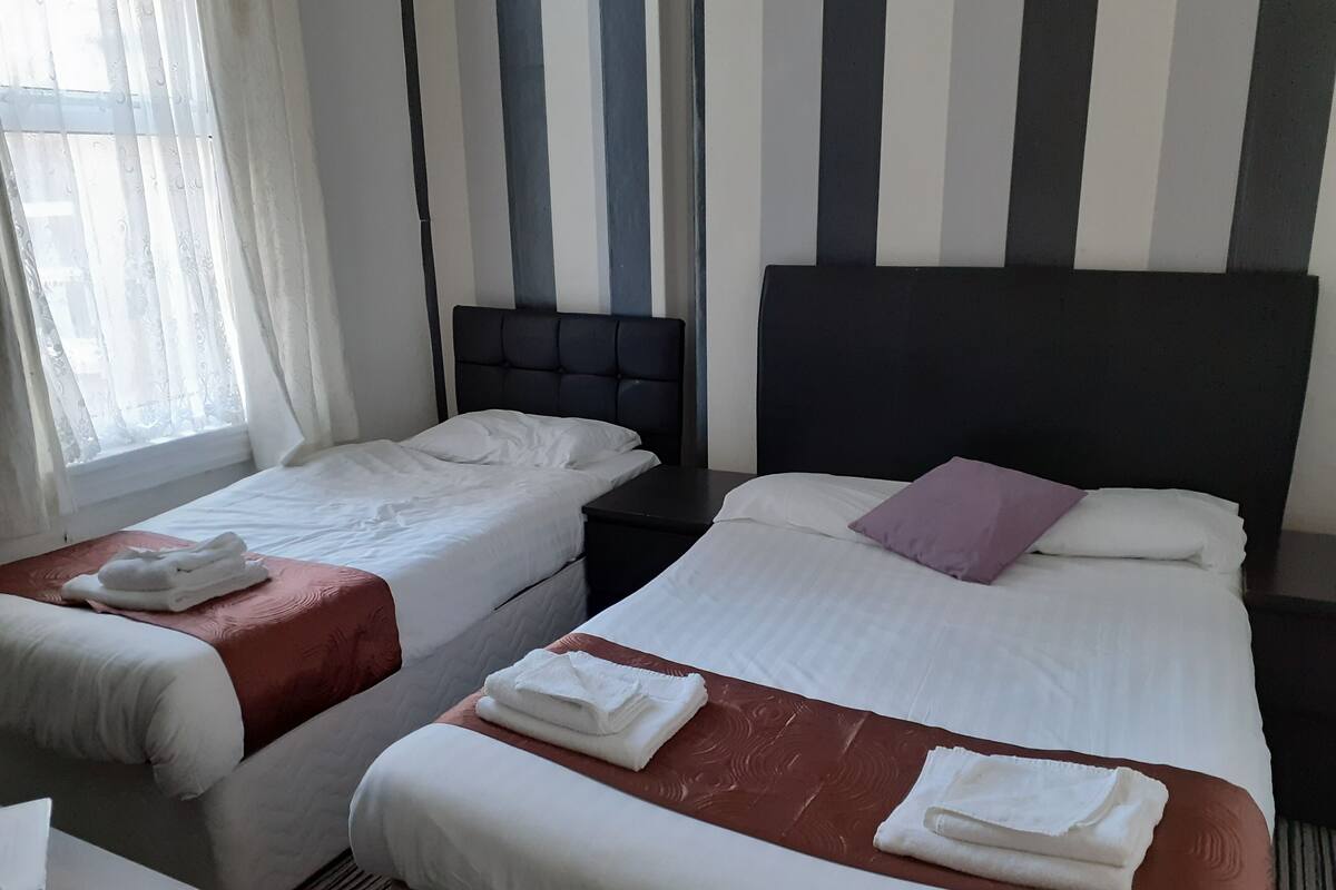 A hotel room with two single beds, each with white linens and towels, a dark headboard, and a window with sheer curtains.