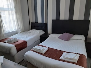 A hotel room with two single beds, each with white linens and towels, a dark headboard, and a window with sheer curtains.