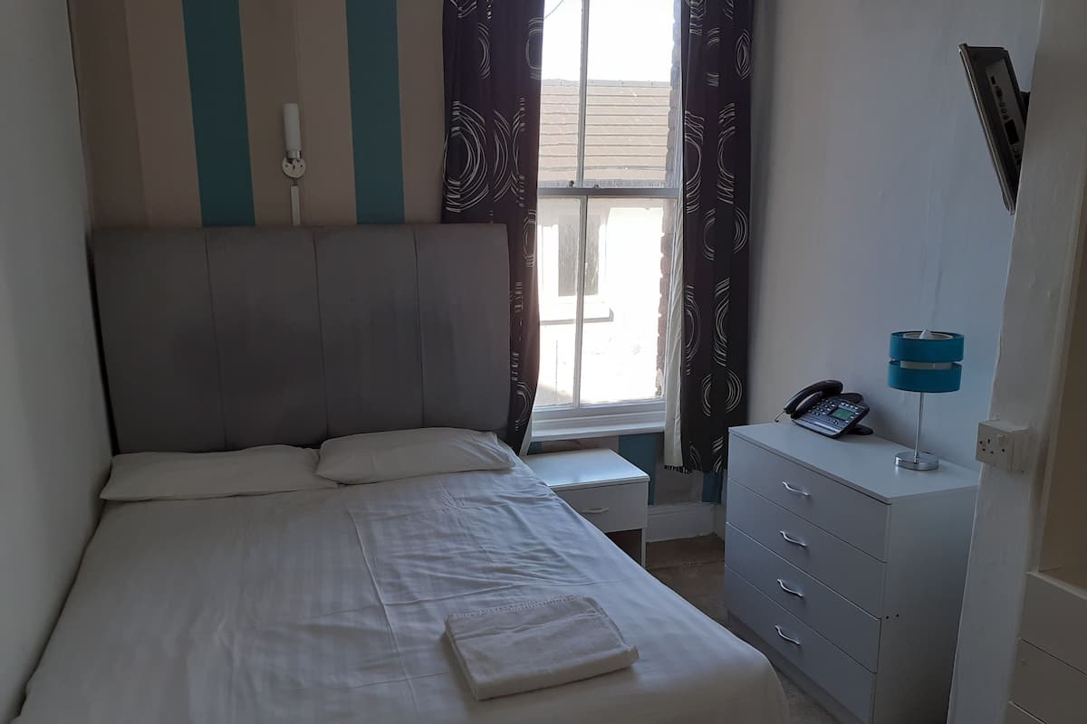 A neatly made bed with pillows, a bedside table with a phone, a lamp, and a window with curtains.