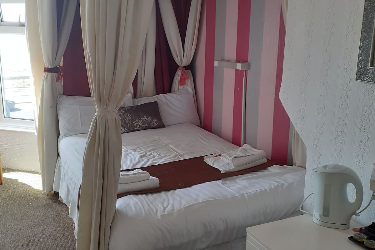 A bedroom with a canopy bed, a bedside table with a kettle, and a dresser.