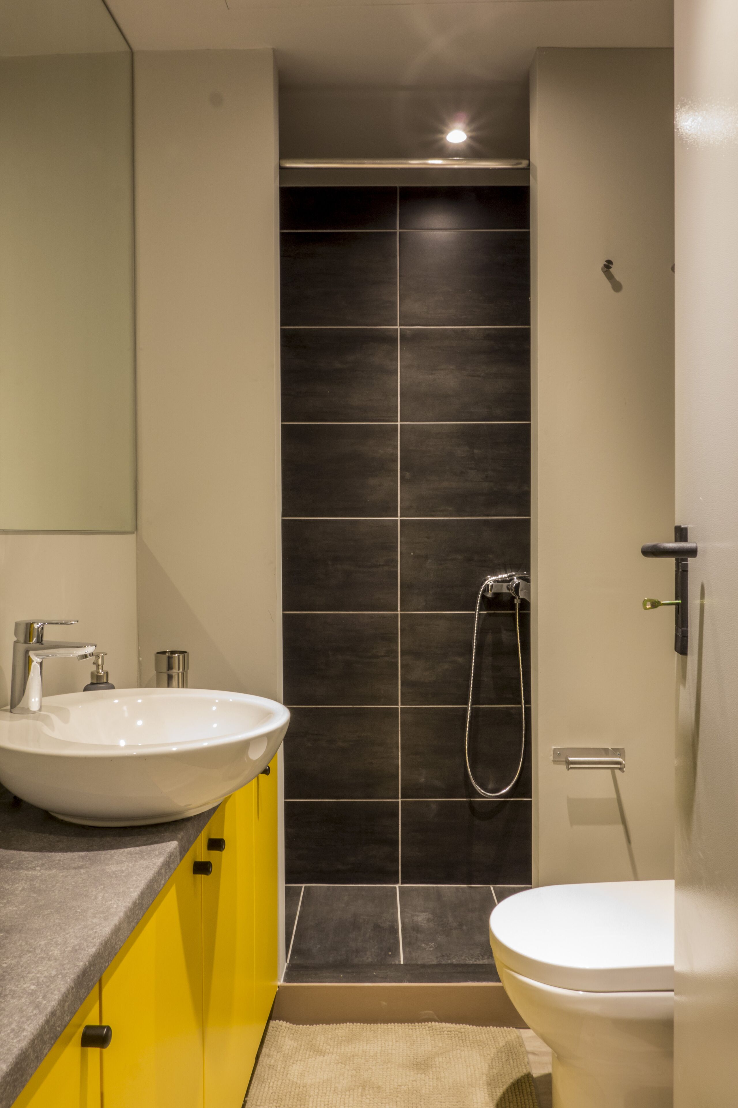 Une salle de bain moderne avec un lavabo blanc, des meubles jaunes, une douche carrelée de couleur sombre et des toilettes blanches.