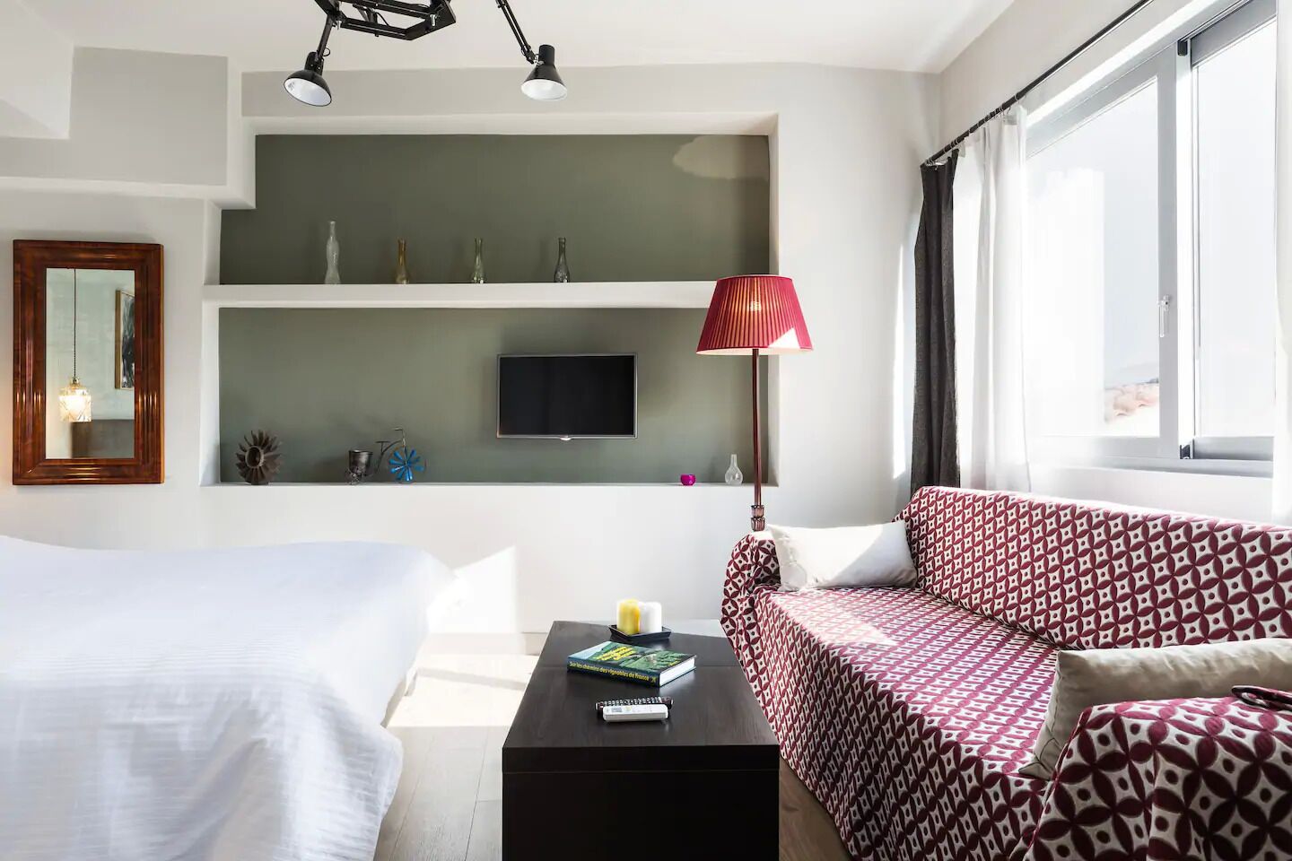 Quarto de hotel com cama, sofá, TV fixada numa parede verde, abajur vermelho e janela com cortinas.