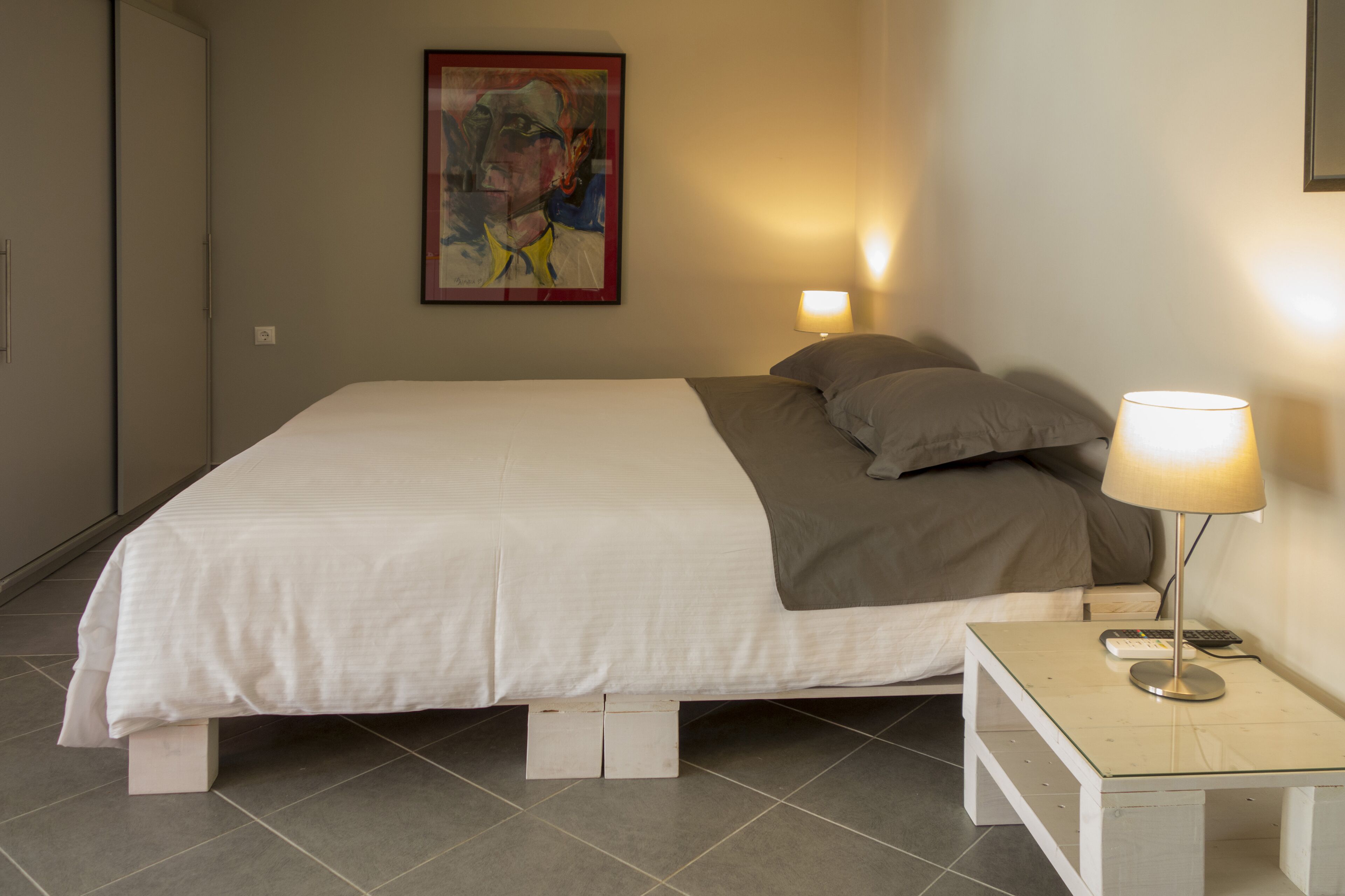 A neatly made bed with a white and gray duvet, a bedside table with a lamp, and a framed abstract painting on the wall.