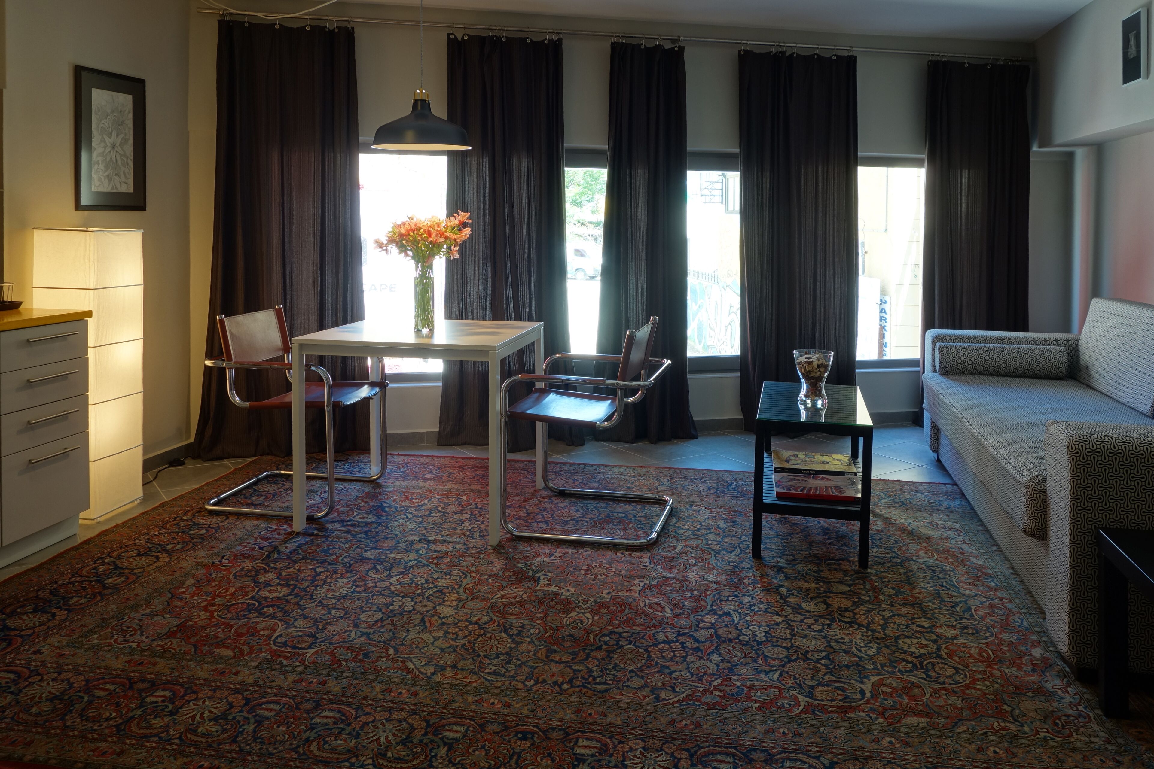 Een woonkamer met een bank, een eettafel met stoelen, een salontafel, een lamp en een groot raam met gordijnen.