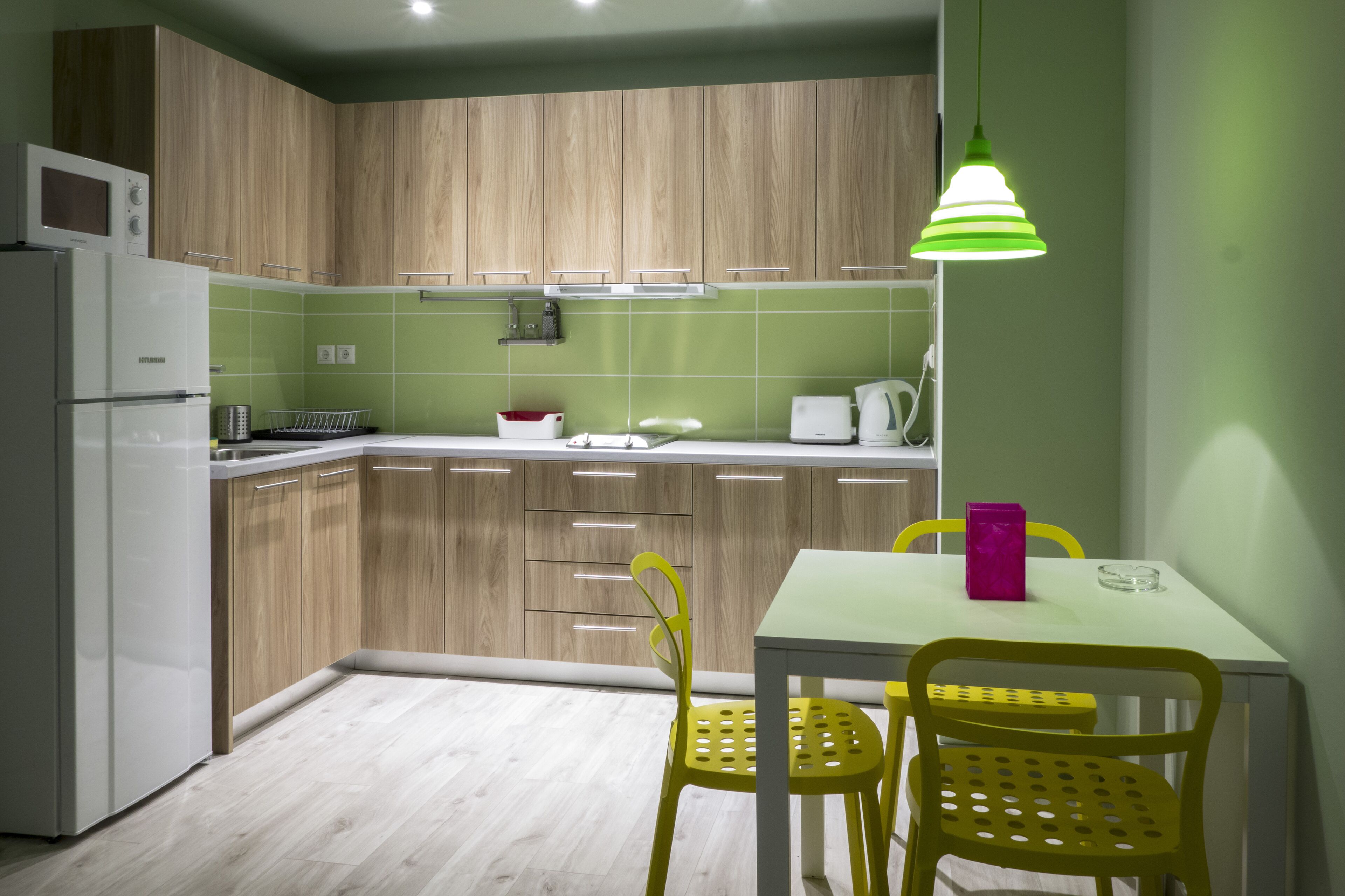 A modern kitchen with wooden cabinets, a white refrigerator, a microwave, and a table with yellow chairs.