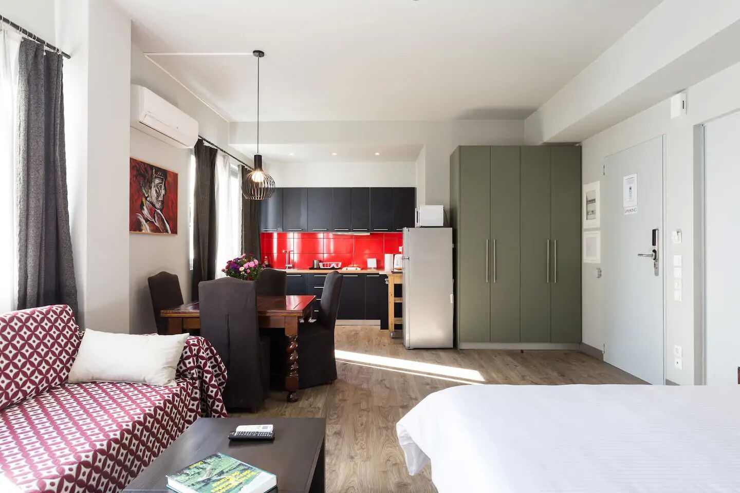 A modern hotel room with a sofa, a bed, a dining table with chairs, a kitchen area with red cabinets, and a green wardrobe.