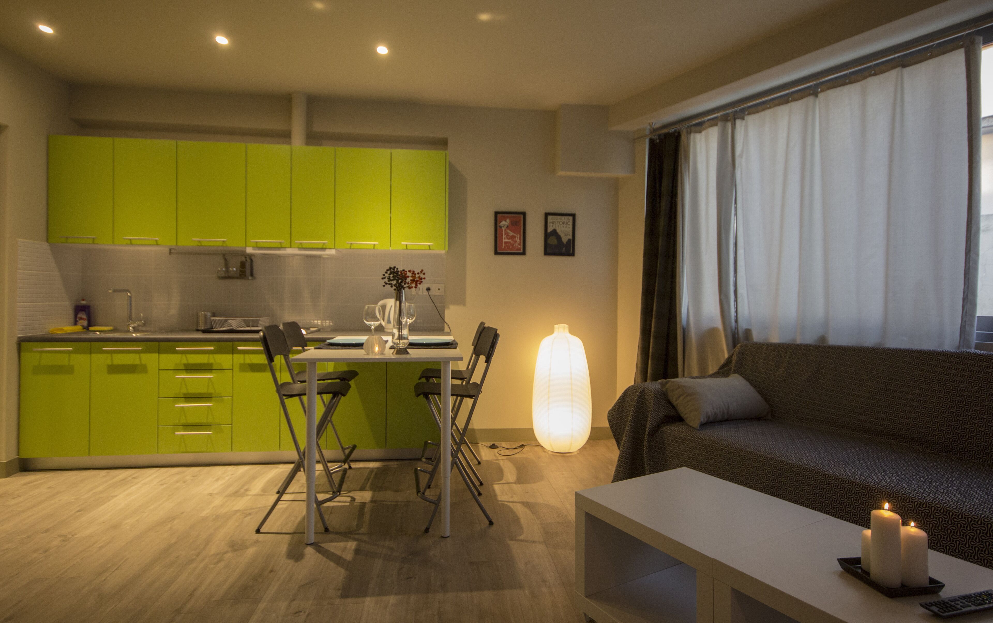A modern kitchen with green cabinets, a dining area with a table and chairs, a sofa, and a large window with curtains.