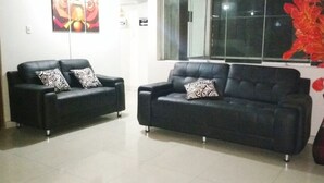 Zwei schwarze Ledersofas mit gemusterten Kissen, ein großes Fenster mit Blick nach draußen und eine rotes Blumenarrangement auf der rechten Seite.