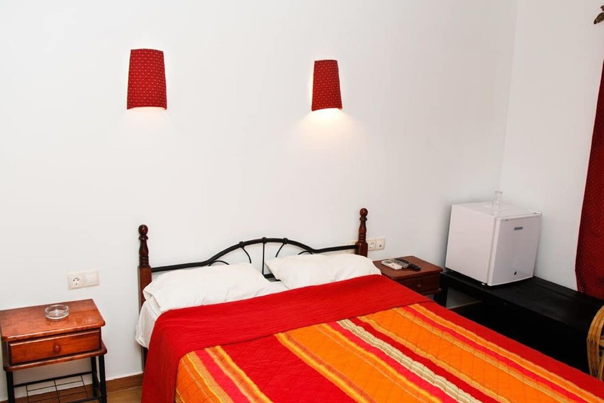 A hotel room with a wooden bed, a bedside table with a lamp, and a small white appliance on the floor.
