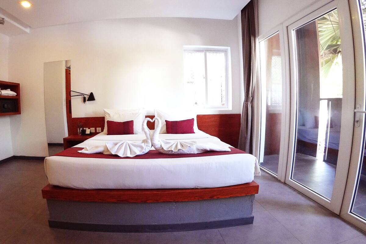 A hotel room with a large bed, a wooden headboard, a red accent wall, a mirror, a wall-mounted lamp, and a sliding glass door leading to a balcony.