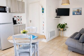 A compact living space with a kitchenette, a dining area with a table and chairs, a gray sofa, a television, and a potted plant. - LE LOGIS, Gîte 1. Clos des Aigrettes, 30m from La Cotinière Port. (Saint-Pierre-d'Oléron)