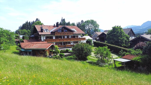 Een groot, meerlaags huis met een rode pannendak, omgeven door weelderige begroeiing en een veld vol gele bloemen.