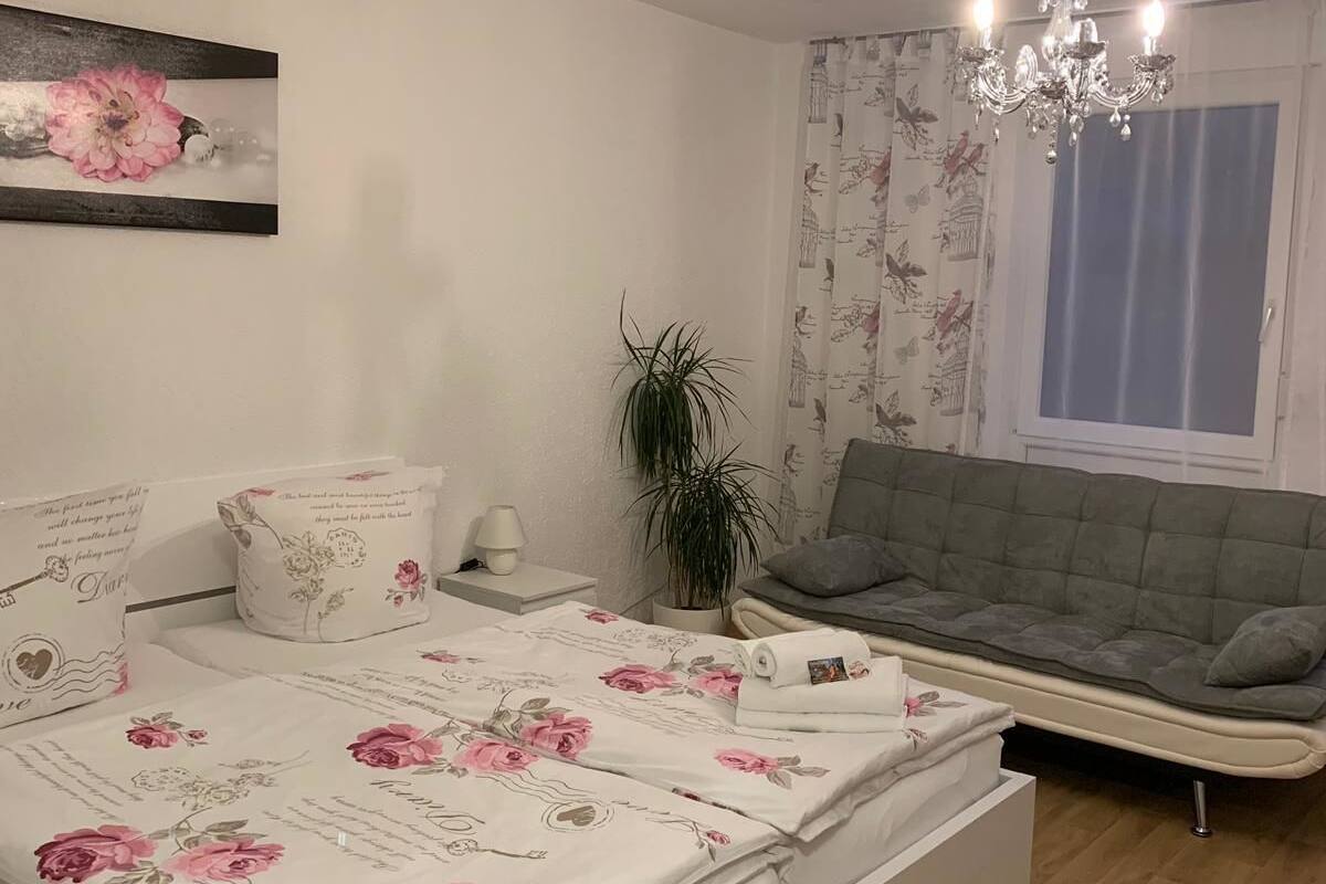 A bedroom with a bed, a grey sofa, a chandelier, and a potted plant.