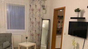 A living room with a sofa, a small table, a mirror, a TV, and a wooden chair. - Pension Nathalie Leser (Baden-Baden)