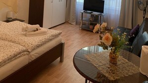 A hotel room with a bed, a desk with a TV, a dining table with a vase of flowers, and a chandelier. - Pension Nathalie Leser (Baden-Baden)