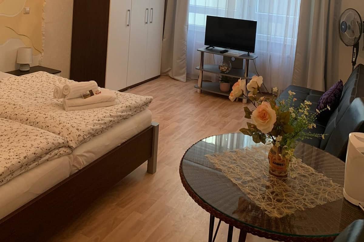 A hotel room with a bed, a desk with a TV, a dining table with a vase of flowers, and a chandelier.