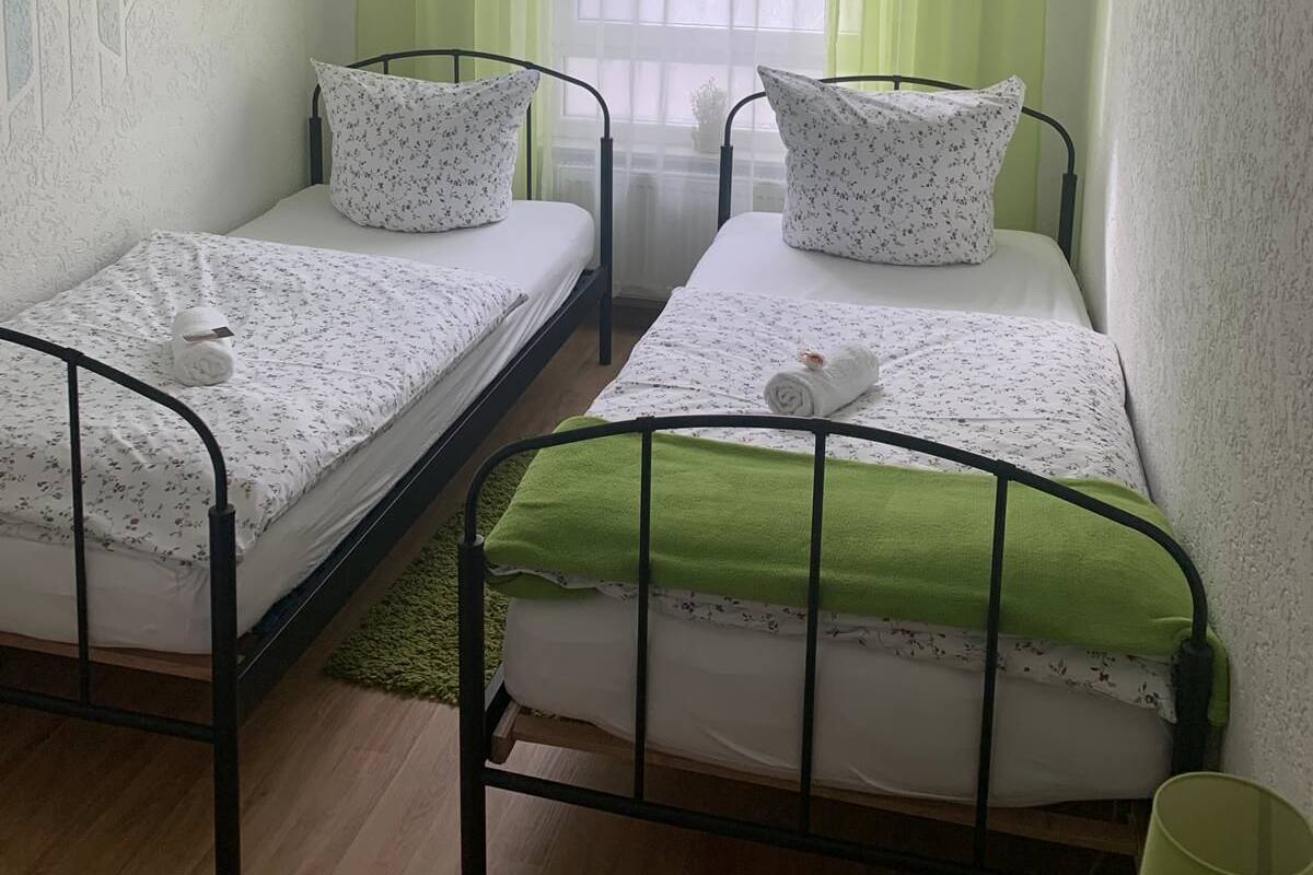 A small, clean room with two single beds, a window with sheer curtains, and a green rug.