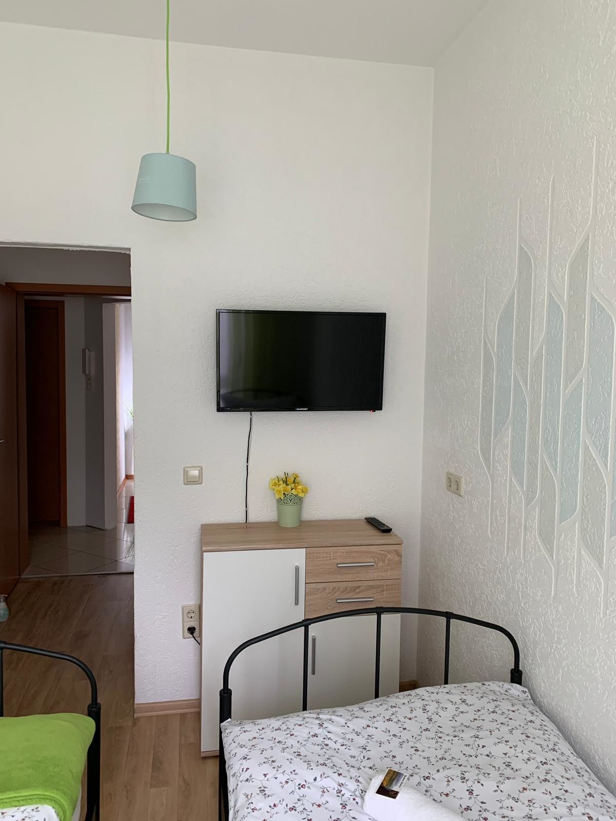 Ein Schlafzimmer mit einem Bett, einem Nachttisch mit Lampe, einem an der Wand montierten Fernseher und einer Tür zu einem anderen Raum.