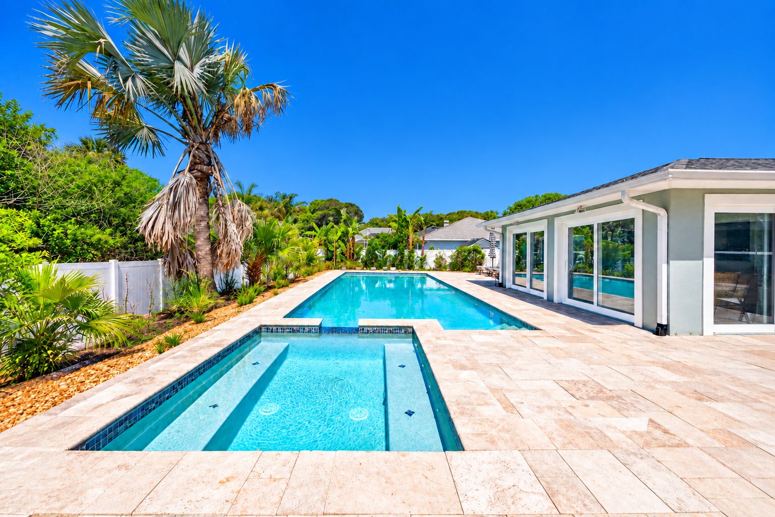 Une piscine rectangulaire bordée d’une terrasse carrelée, entourée de palmiers et d’une maison moderne dotée de grandes fenêtres.