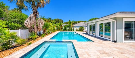 A rectangular swimming pool with a tiled deck, surrounded by palm trees and a modern house with large windows.