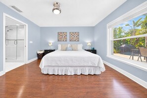 A bedroom with a large bed, a glass shower enclosure, two bedside tables with lamps, and a window with a view of greenery.