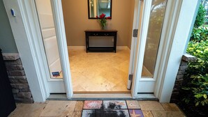 Une porte qui donne sur une pièce dont le sol est recouvert de carreaux, où l’on trouve une petite console sur laquelle est posé un vase de fleurs, et un miroir accroché au mur.