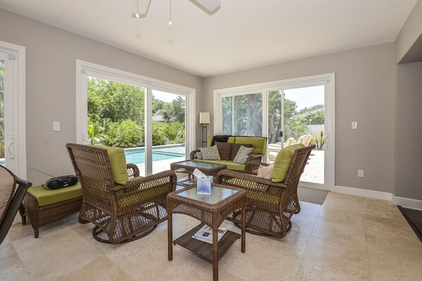 A spacious room with a glass-top table surrounded by wicker chairs and a sofa, large windows offering a view of a swimming pool and greenery. - Whispering Palms - Beach-Side Pool Home (Ponce Inlet)