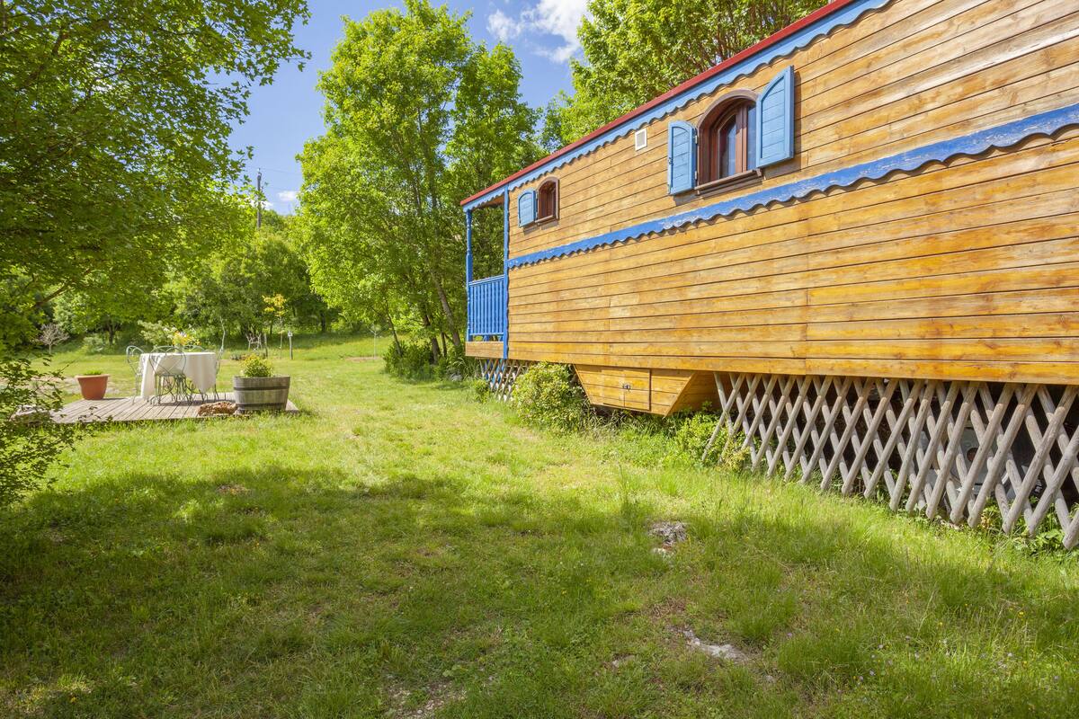A wooden house with a red roof and blue shutters, surrounded by a wooden fence and lush greenery.