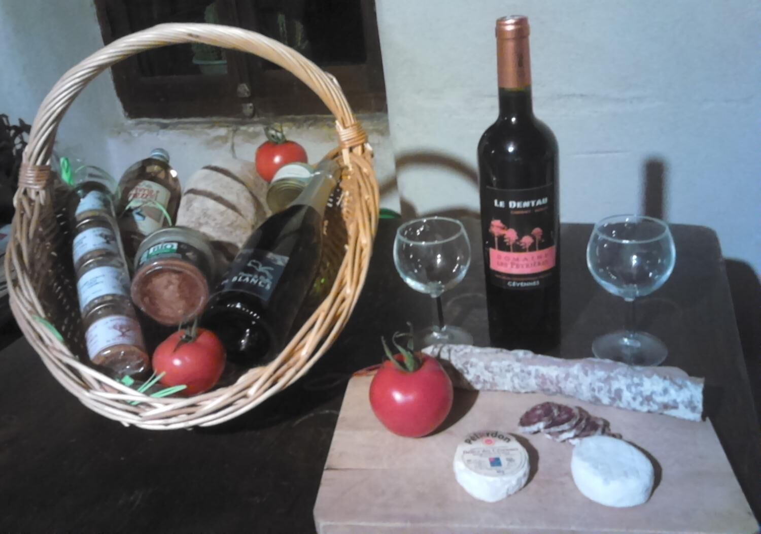Una mesa con una botella de vino, dos copas, una cesta de comida y una tabla de madera con queso y tomate.