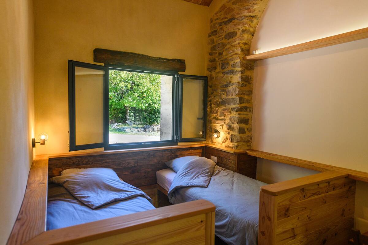 A cozy room with two wooden beds, a stone wall, and a window with a view of greenery.