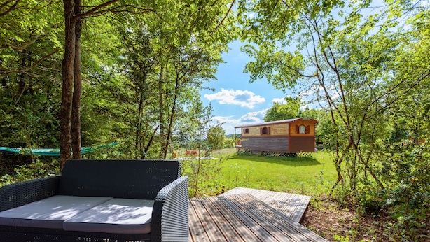 A wooden deck with a wicker sofa and a small wooden structure in a forested area. - La Grange d'Emile Gites, Roulottes et Piscine (Blandas)