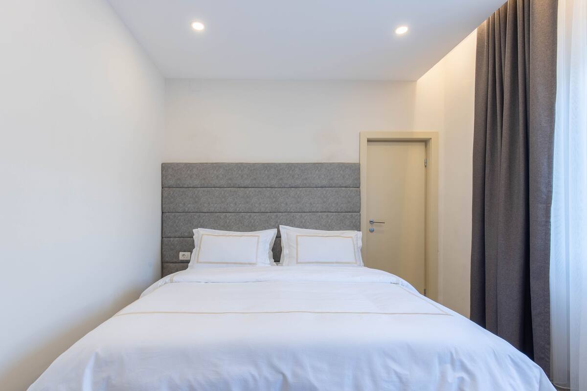 A modern hotel room with a large bed, a grey headboard, and a door to the right.