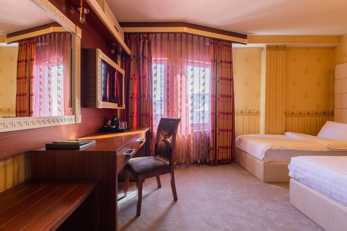 A hotel room with a desk, chair, two beds, and large windows with curtains.