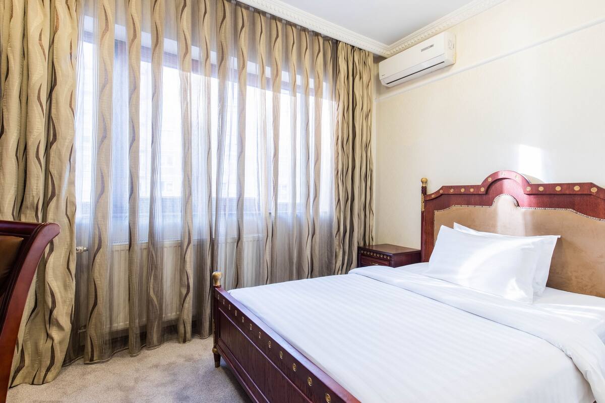 A hotel room with a bed, curtains, an air conditioning unit, and a nightstand.